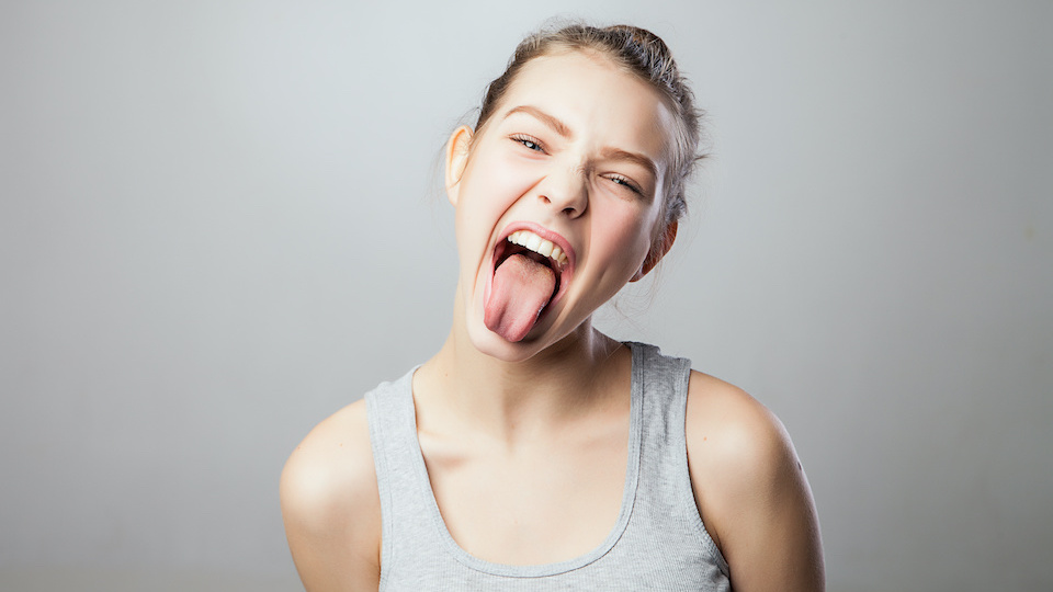 Young pretty woman shoots out tongue, isolated on grey. 